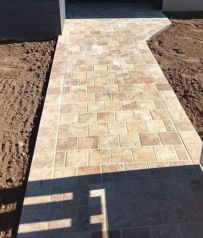 Une allée pavée de pierre beige mène à l'entrée d'une maison, bordée de zones de terre nue.