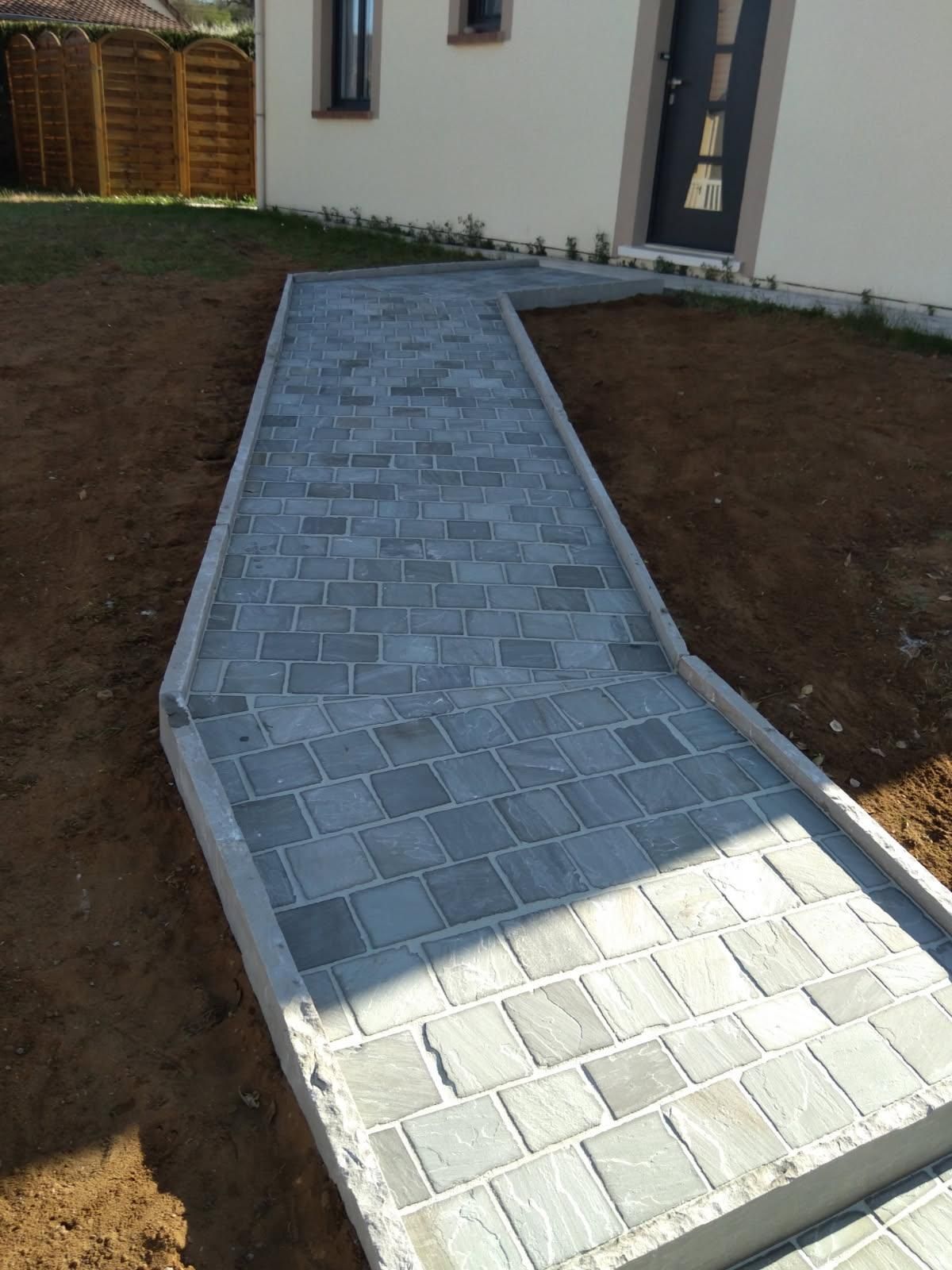 Une allée en pierre grise nouvellement pavée mène à la porte d'entrée d'une maison aux tons clairs, bordée de terre nue.