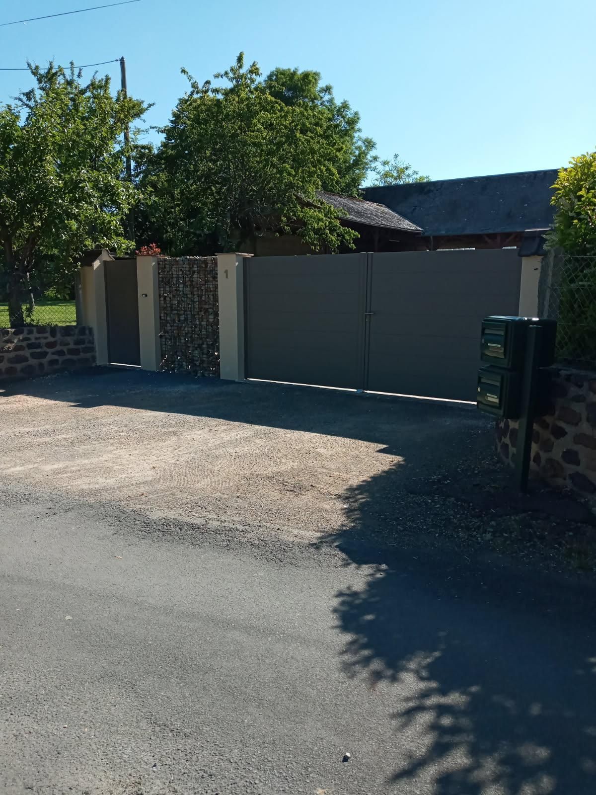 Une allée de gravier menant à un portail en métal gris encastré dans un mur de maçonnerie, bordée d'arbres et d'une boîte aux lettres verte.