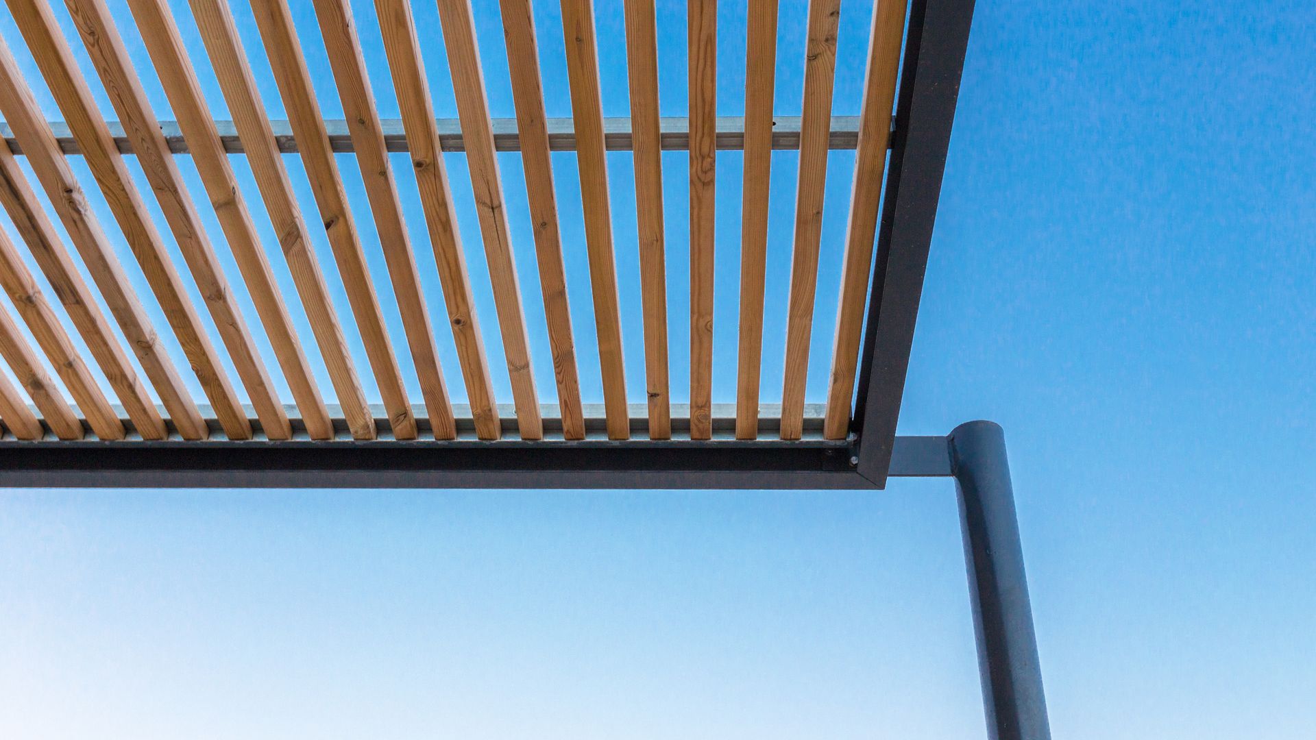 Un toit de pergola en bois à lames parallèles se détachant sur un ciel bleu clair, soutenu par une structure et un pilier en métal noir.