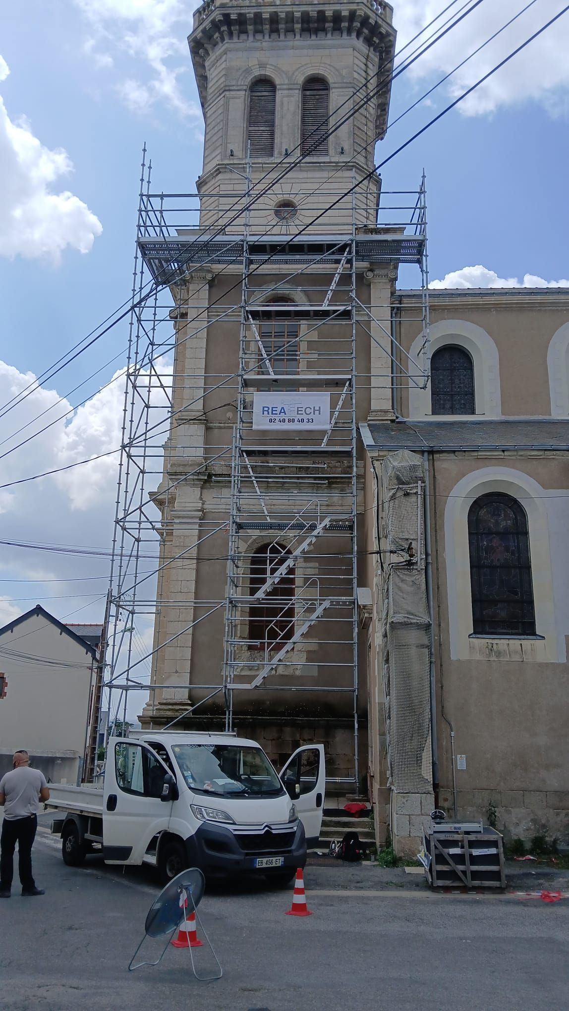 Le clocher en pierre de l'église de Savenay, entouré d'échafaudages, et un camion utilitaire blanc garé devant.