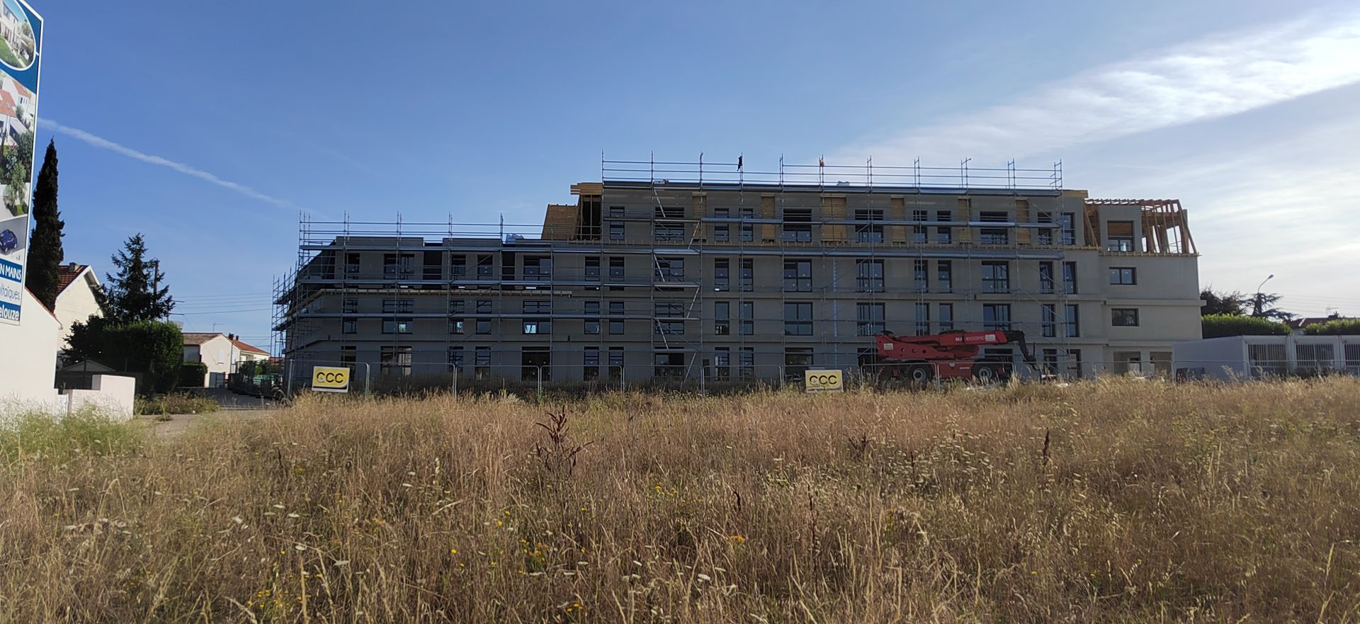 Bâtiment long avec échafaudages, un champ herbeux en premier plan.
