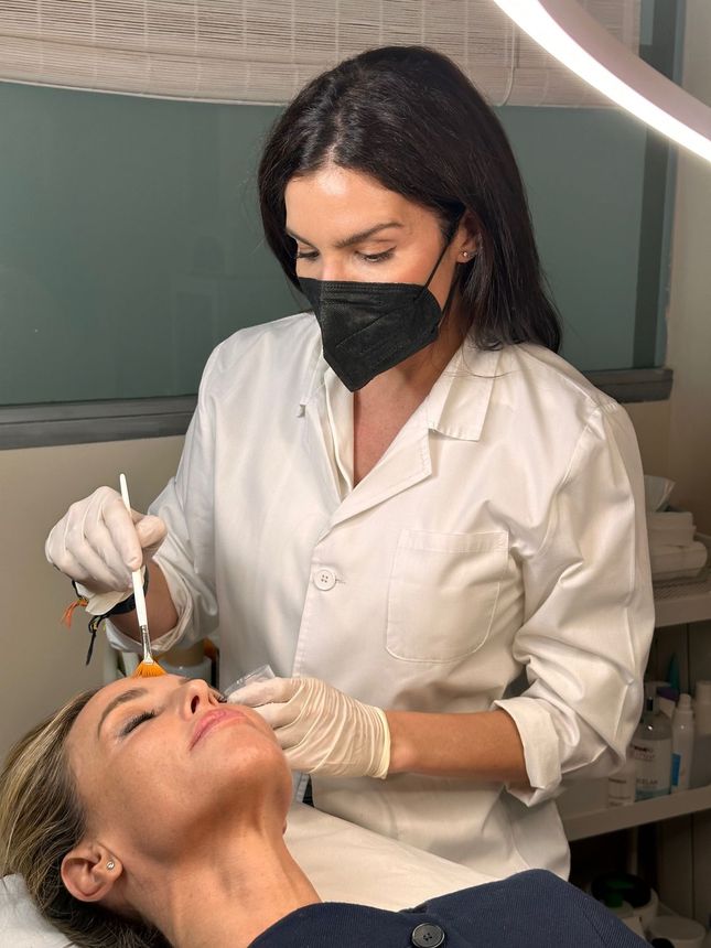 Una mujer que lleva una máscara le está dando a otra mujer un tratamiento facial.