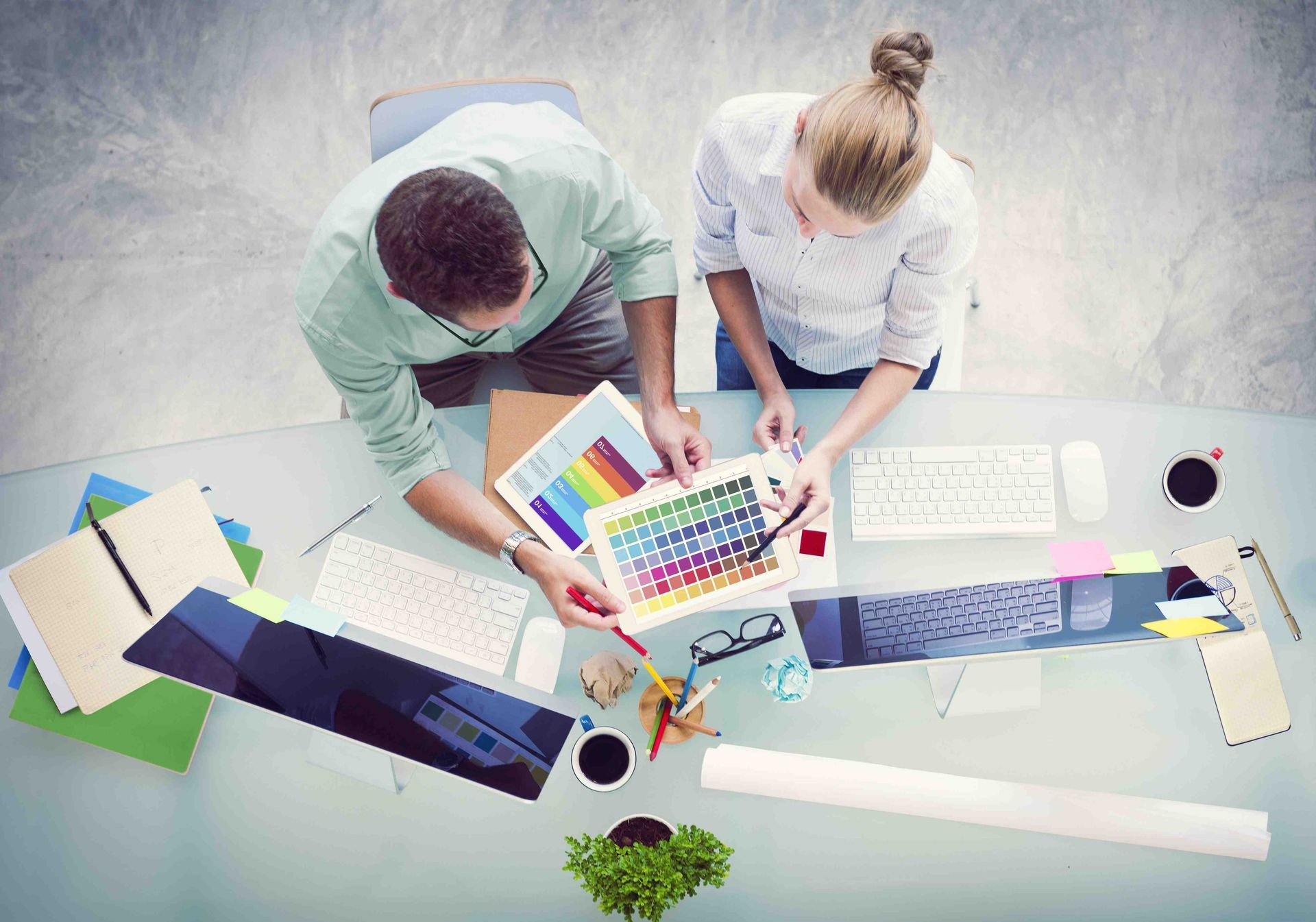 Dos personas en un escritorio colaboran, mirando muestras de color y datos. Se ven teclados de computadora y tazas de café.