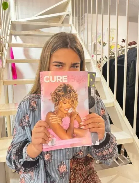 Una persona sostiene una revista titulada “CURE” frente a su rostro en una escalera interior.
