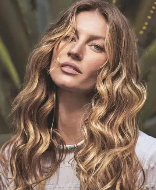 Mujer con cabello rubio ondulado y expresión neutra, vestida con una blusa blanca bajo una iluminación interior tenue.