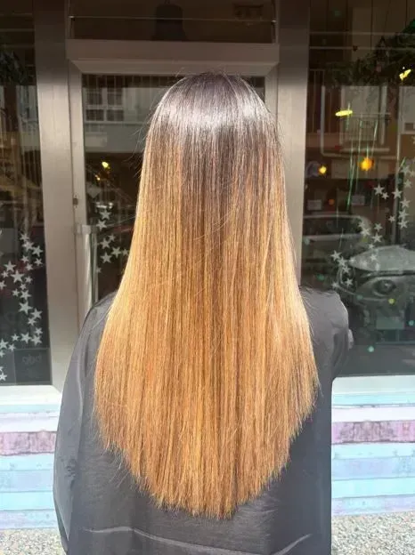 Persona con cabello largo y liso, de color castaño a rubio, con mechas degradadas, de pie al aire libre frente a un edificio.