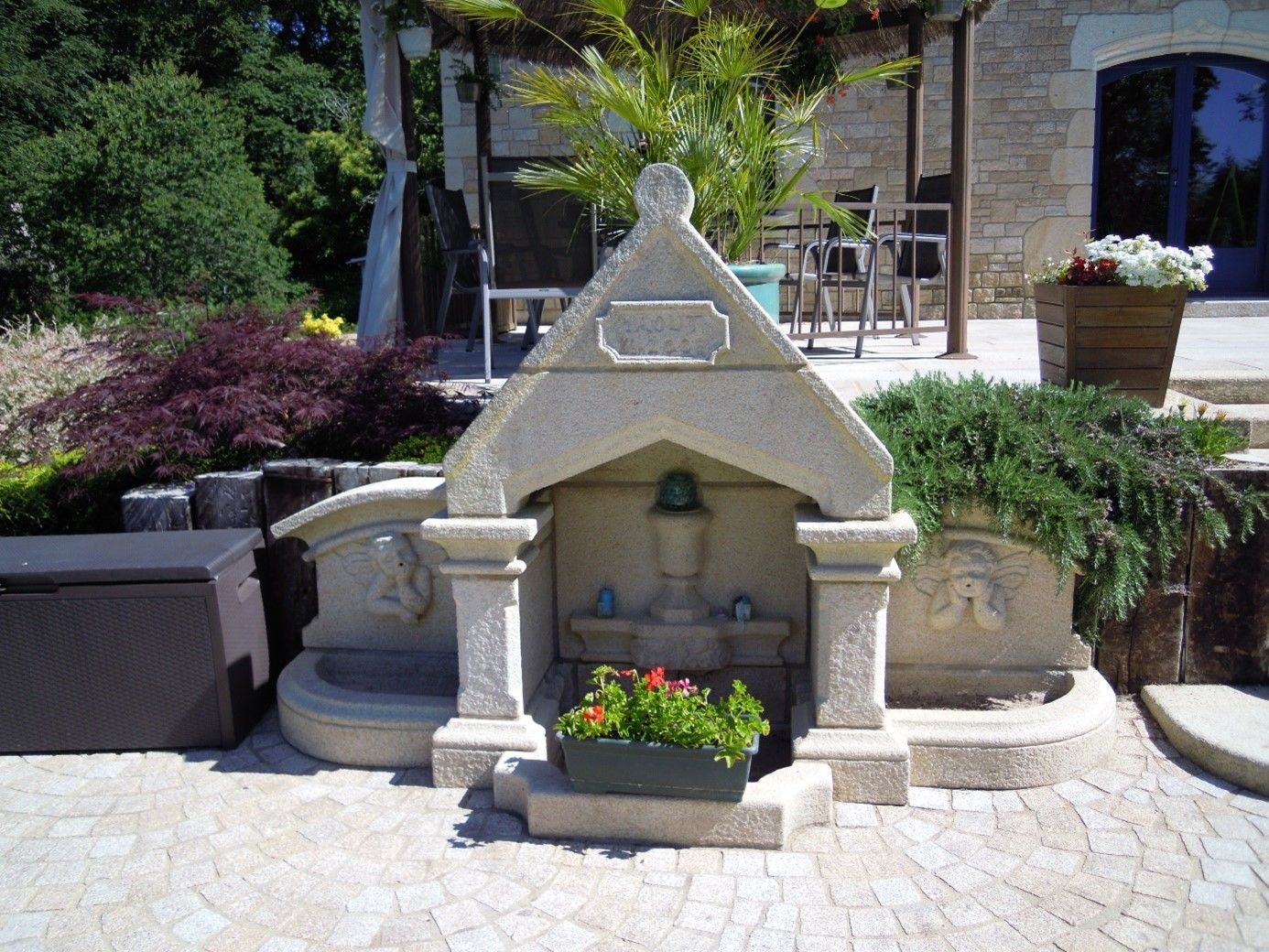 3 fontaines en pierre, celle du milieu possède un toit en pointe et un pot de fleurs devant.