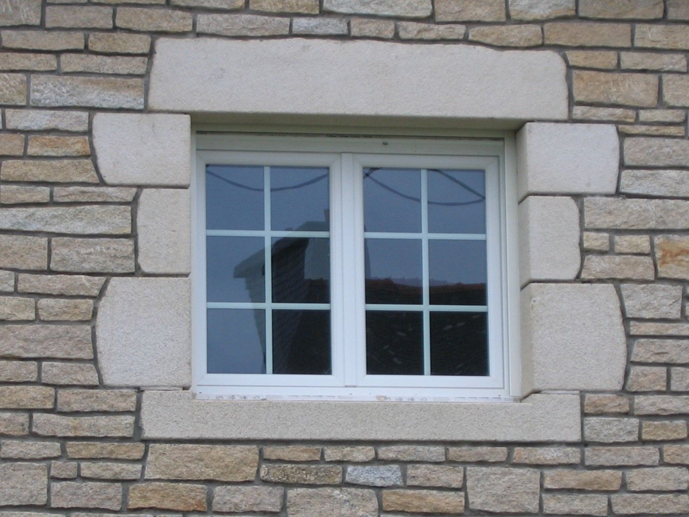 Encadrement en pierres posées horizontalement pour entourer une fenêtre blanche.