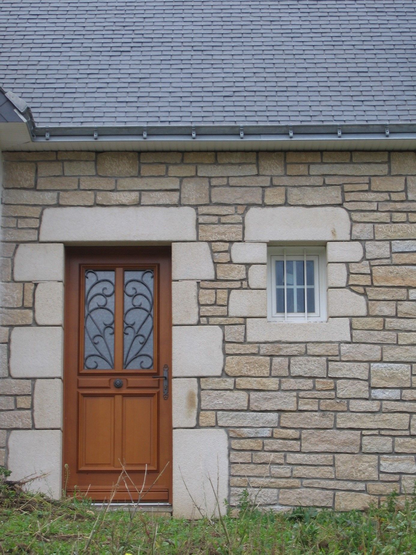 Encadrement en pierre d'une petite fenêtre blanche et d'une porte d'entrée en bois.