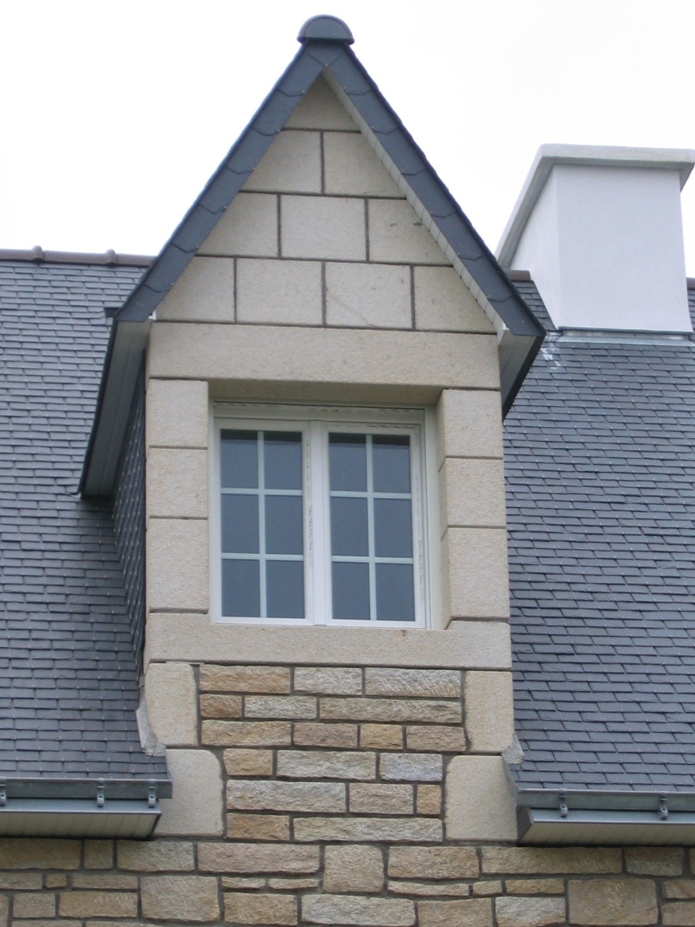 Encadrement en pierre clair d'une fenêtre blanche située sous une toiture.
