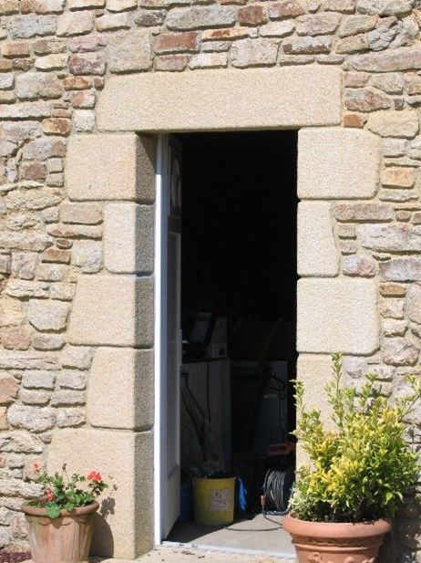 Porte d'entrée blanche avec un encadrement en pierre et deux pots de fleurs de chaque côté.
