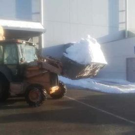 Una pala cargadora transporta una gran pila de nieve junto a un edificio blanco en un estacionamiento.