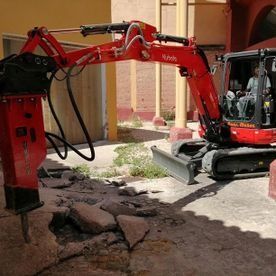 Una excavadora roja demuele el pavimento agrietado en un patio junto a un edificio.