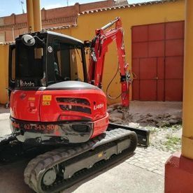 Miniexcavadora roja estacionada junto a un muro amarillo y una puerta roja en un patio pavimentado.
