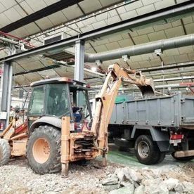 Retroexcavadora junto a un camión volquete gris dentro de un almacén industrial, con escombros en el suelo.