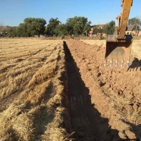 Excavadora cavando una zanja junto a un campo cosechado bajo un cielo despejado.