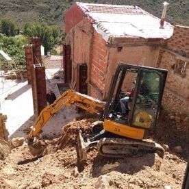 Una excavadora amarilla está removiendo tierra junto a una casa de ladrillos dañada en una ladera empinada.