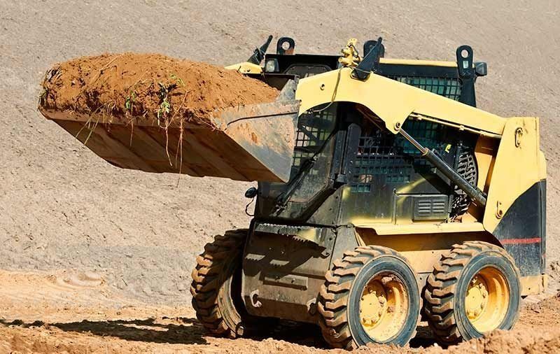 Minicargadora amarilla transportando tierra con la pala elevada en una obra de construcción.