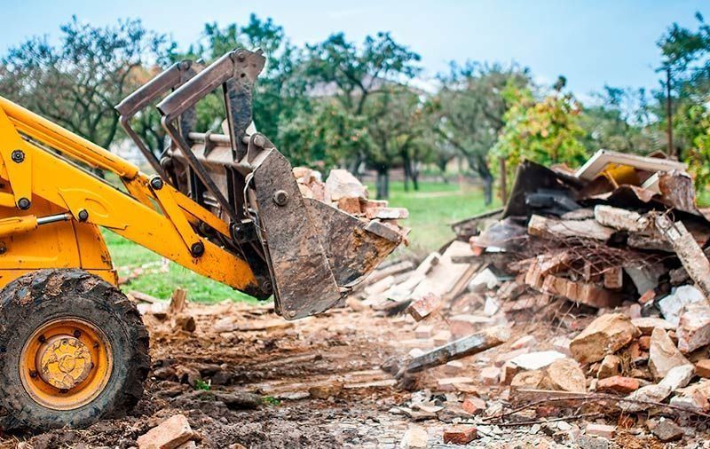 Una excavadora amarilla demuele un montón de escombros al aire libre con árboles al fondo.