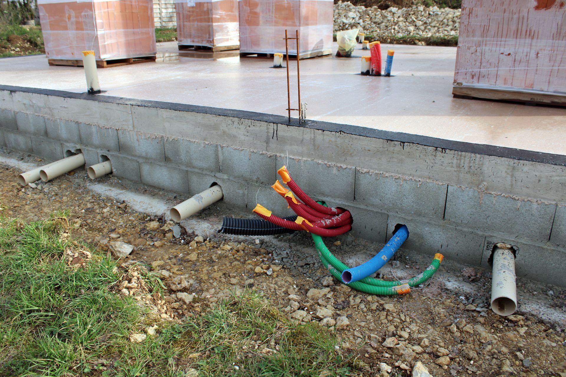 Câbles et tuyaux colorés apparents près d'un mur de fondation en béton.