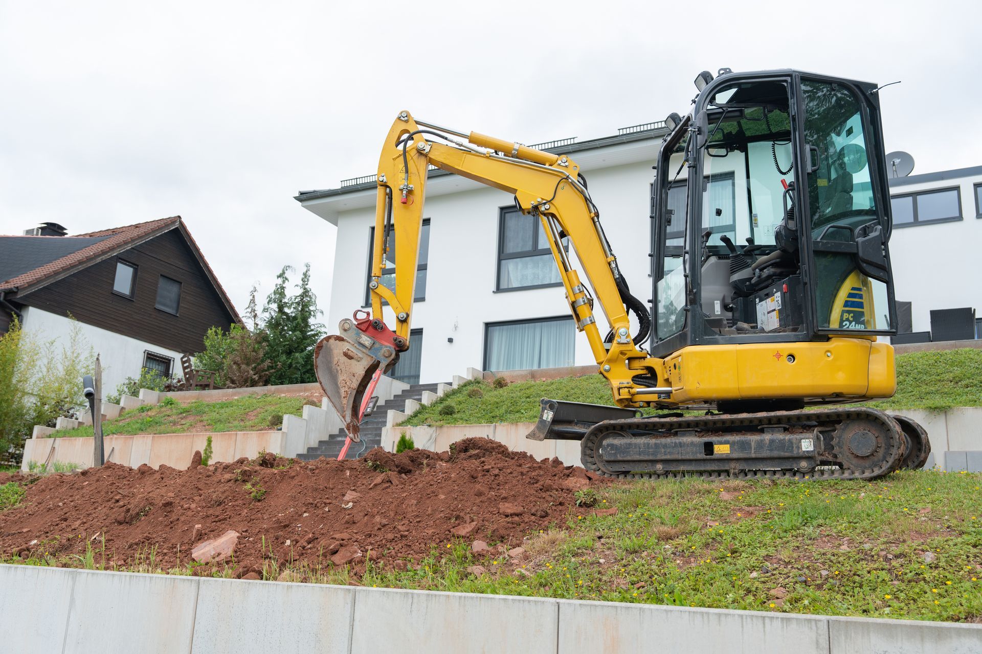 Une pelleteuse jaune creuse la terre à côté d'une maison contemporaine sur une pente herbeuse.