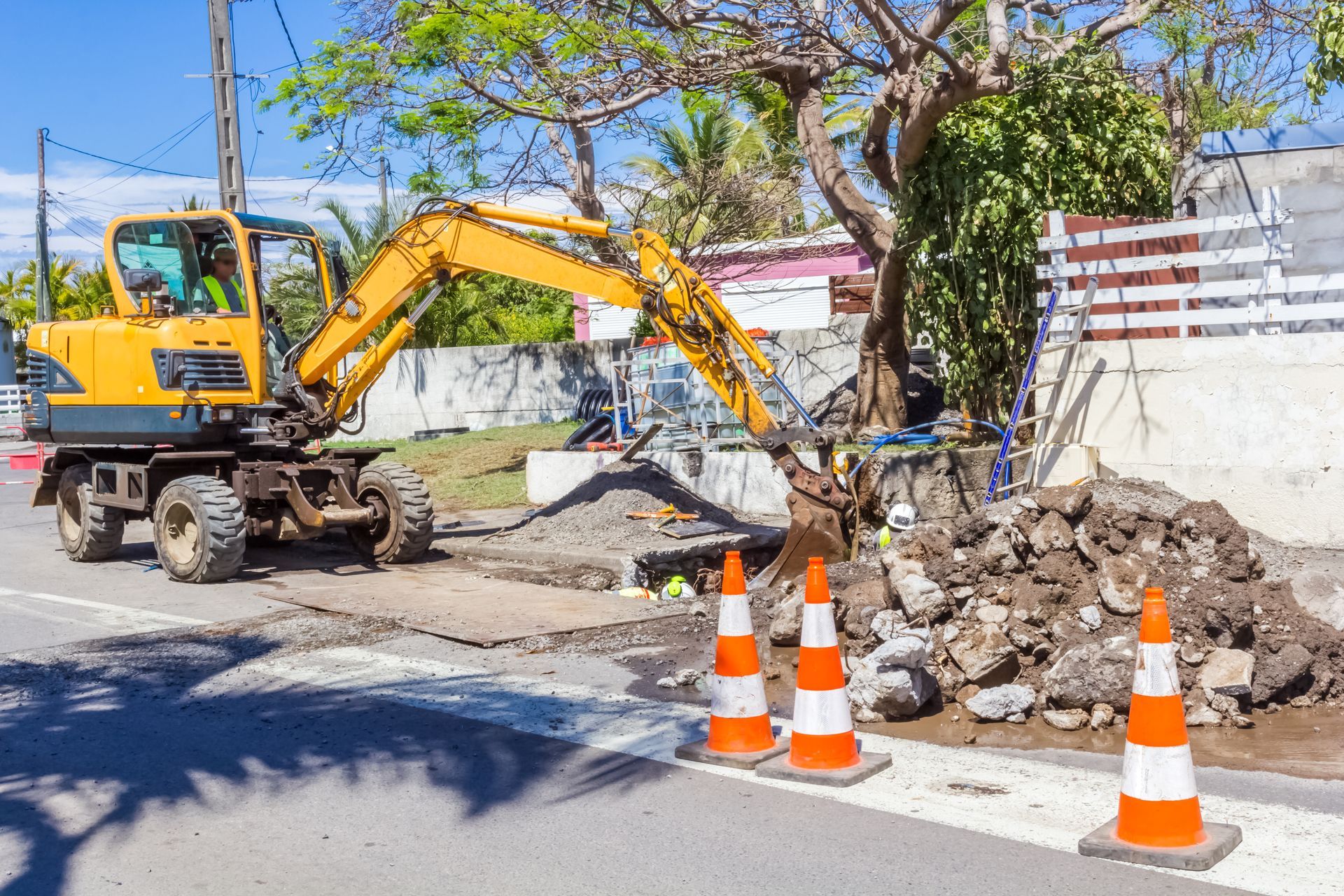 Une pelleteuse jaune creuse des gravats à côté de cônes de signalisation dans une rue bordée d'arbres.