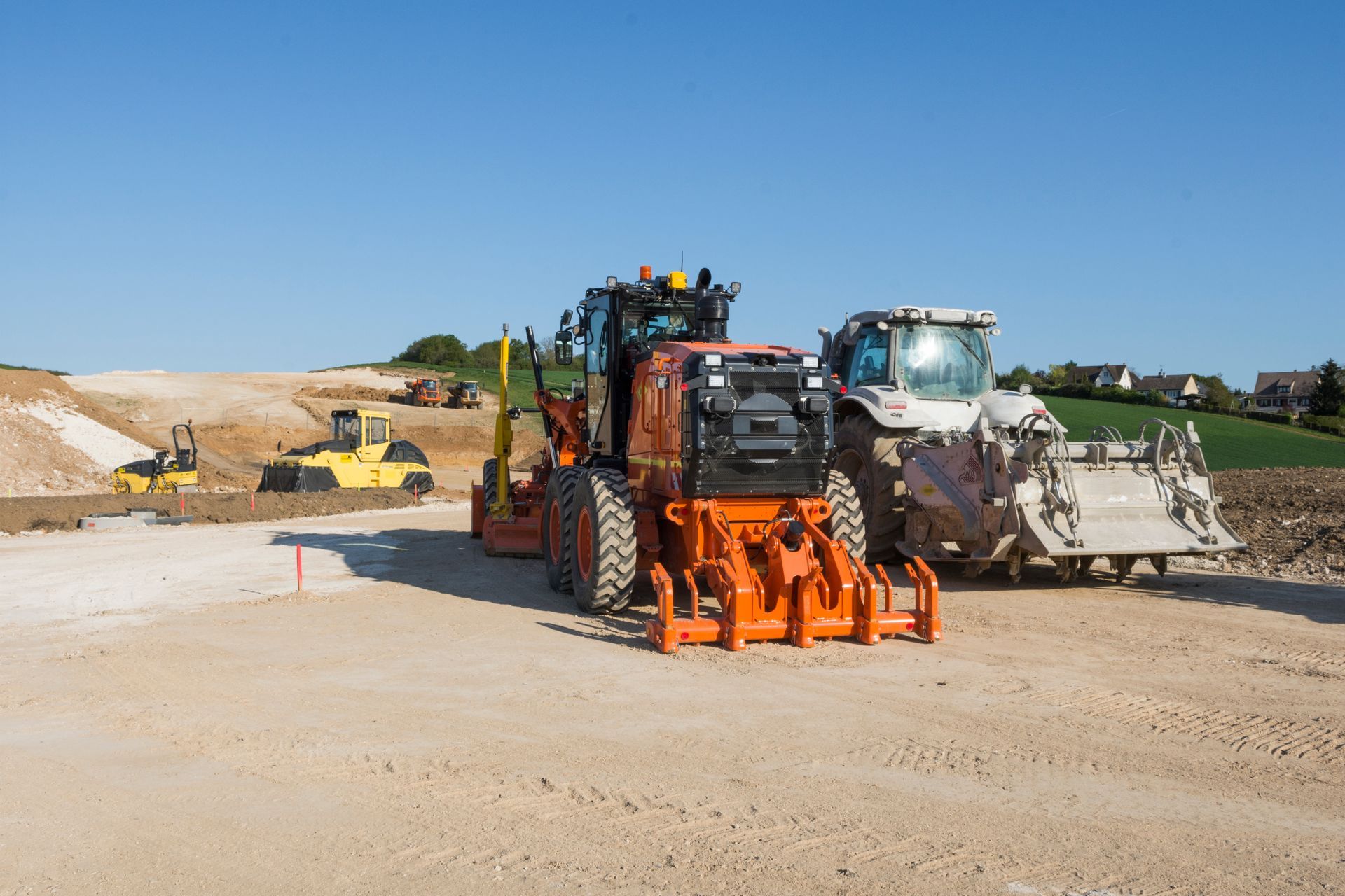 Plusieurs engins de travaux publics, dont une niveleuse orange, une stabilisatrice blanche et des compacteurs, sur un chantier de terrassement.