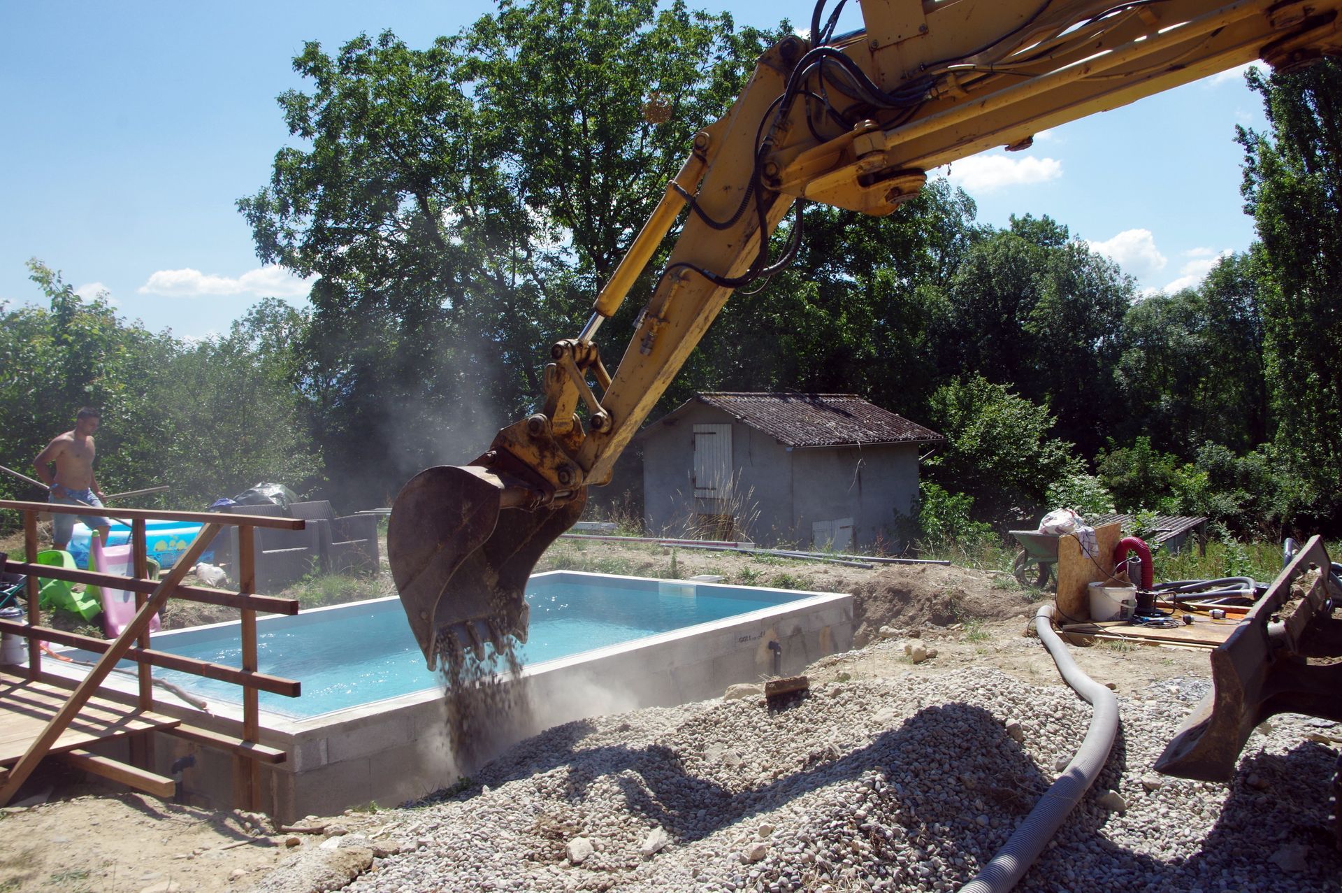 Une pelleteuse déverse des cailloux et de la terre à côté d'une piscine en construction, pendant des travaux de terrassement.