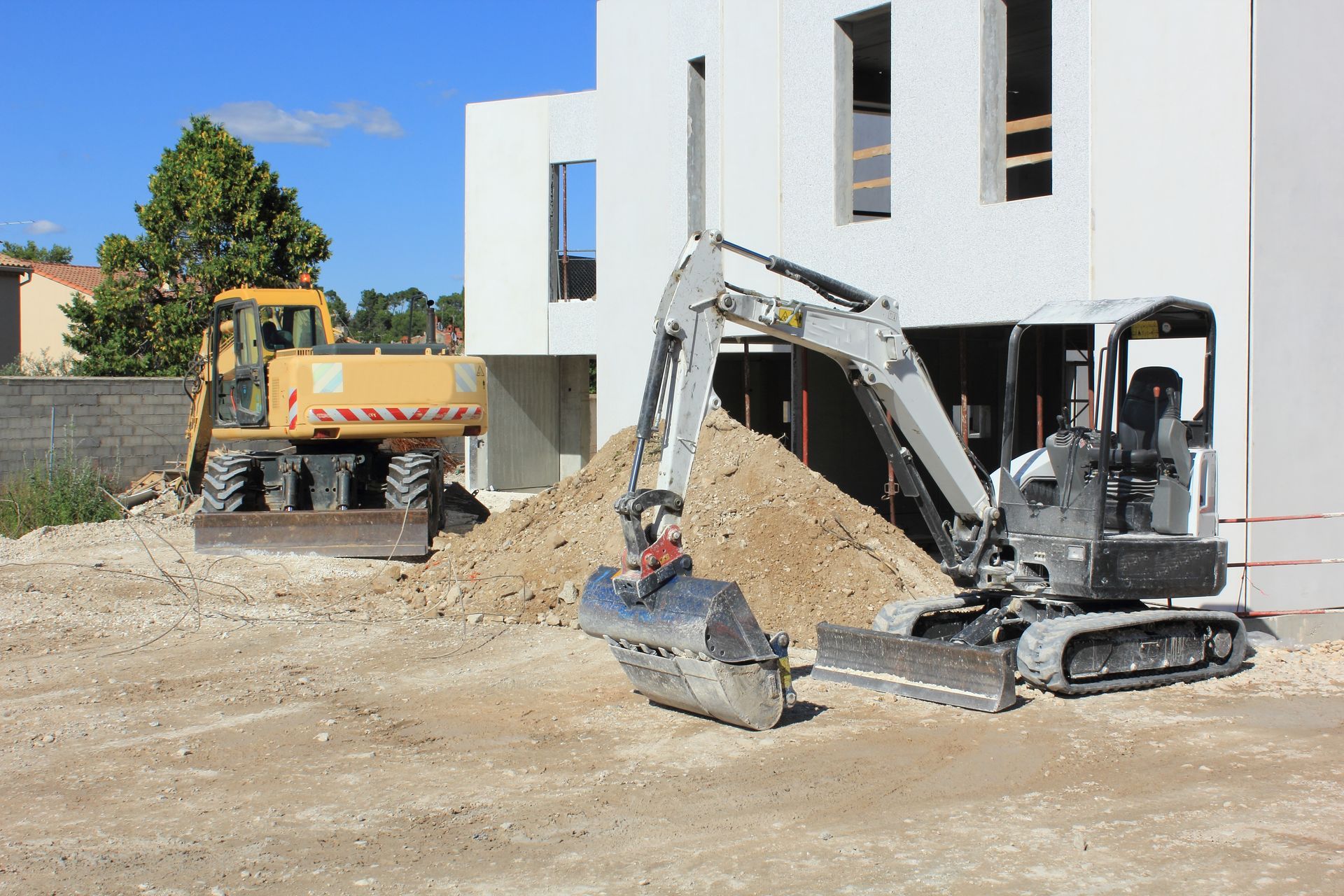 Une excavatrice et un camion-benne sur un chantier de construction, à côté d'un bâtiment blanc.