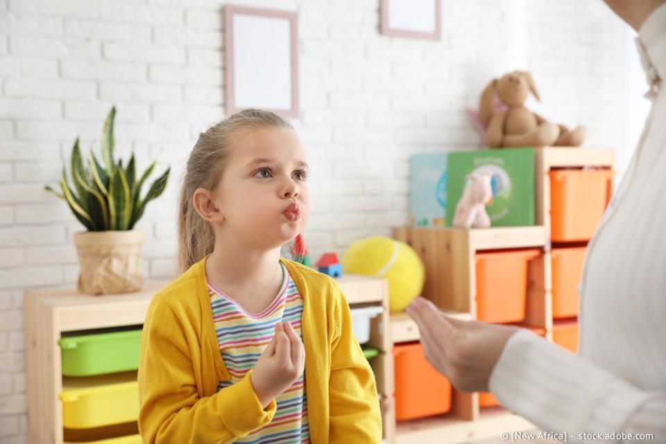 Mädchen in gelber Strickjacke übt mit Erwachsenen in einem Spielzimmer das Sprechen.