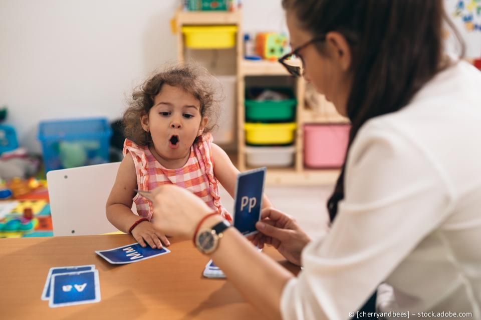 Ein Kind schaut mit überraschtem Gesichtsausdruck auf eine Karte mit dem Buchstaben „Pp“, die ein Erwachsener an einem Tisch hält.