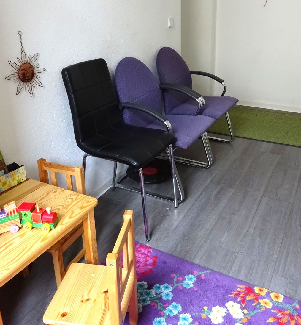 Ein Wartezimmer mit Stühlen in verschiedenen Farben, einem Tisch mit Spielzeug und einem Teppich.