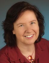 Frau mit dunkelbraunem Haar lächelt, trägt eine rote Bluse und eine rote Halskette, vor blauem Hintergrund.