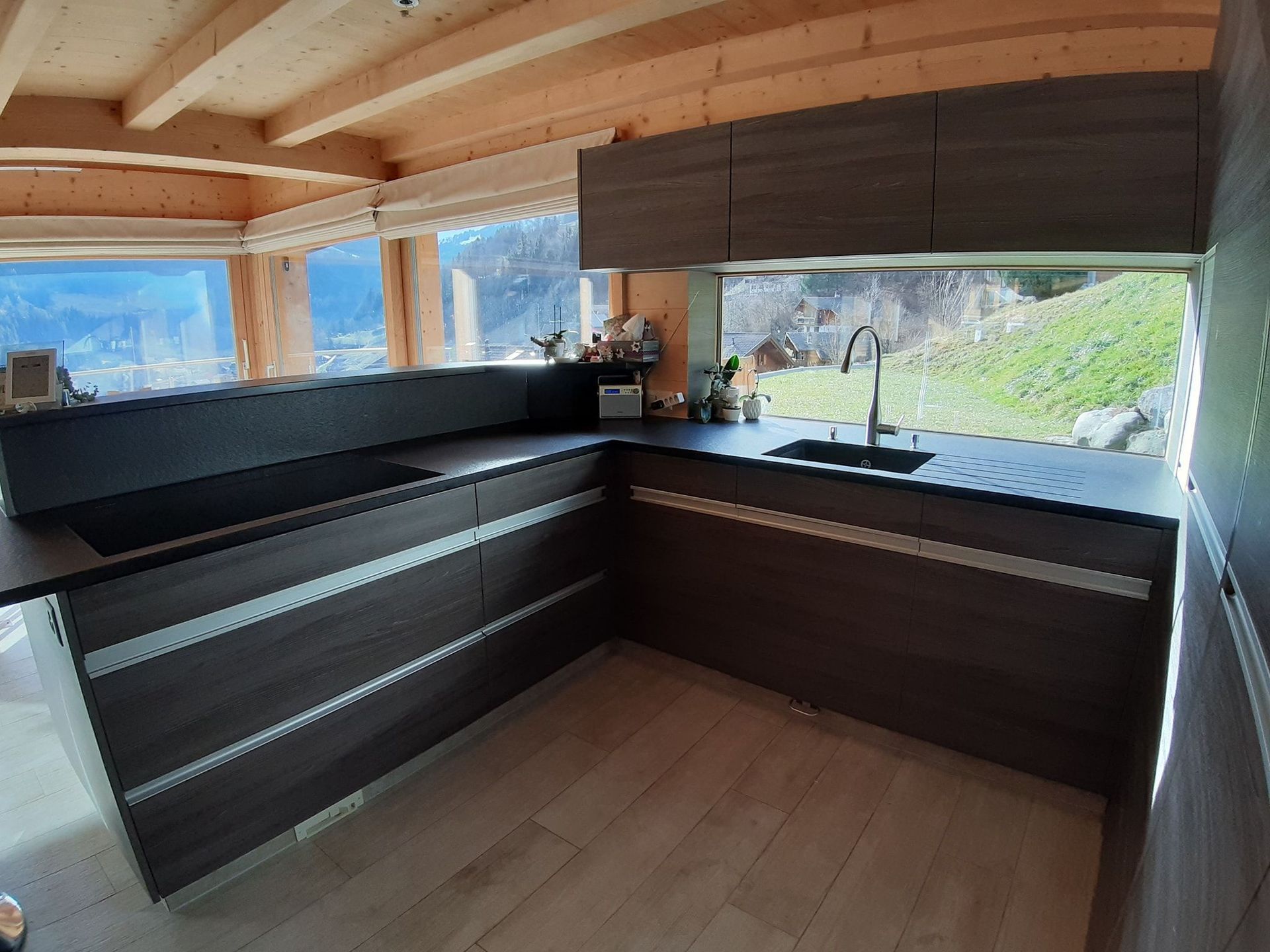 Cuisine en stratifiée gris imitation bois, plan de travail en granit noir absolu, poignées profilées en alu, hotte de ventilation intégrée dans le plan de travail, store déroulant et porte secrète qui donne sur le cellier!