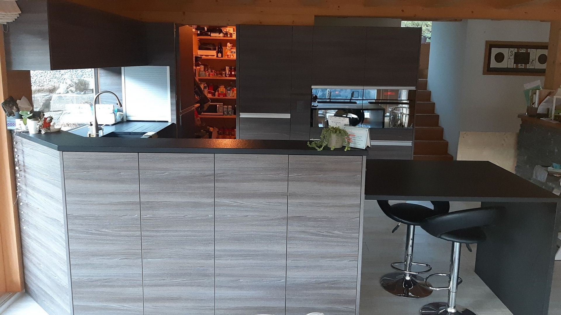 Cuisine en stratifiée gris imitation bois, plan de travail en granit noir absolu, poignées profilées en alu, hotte de ventilation intégrée dans le plan de travail, store déroulant et porte secrète qui donne sur le cellier!