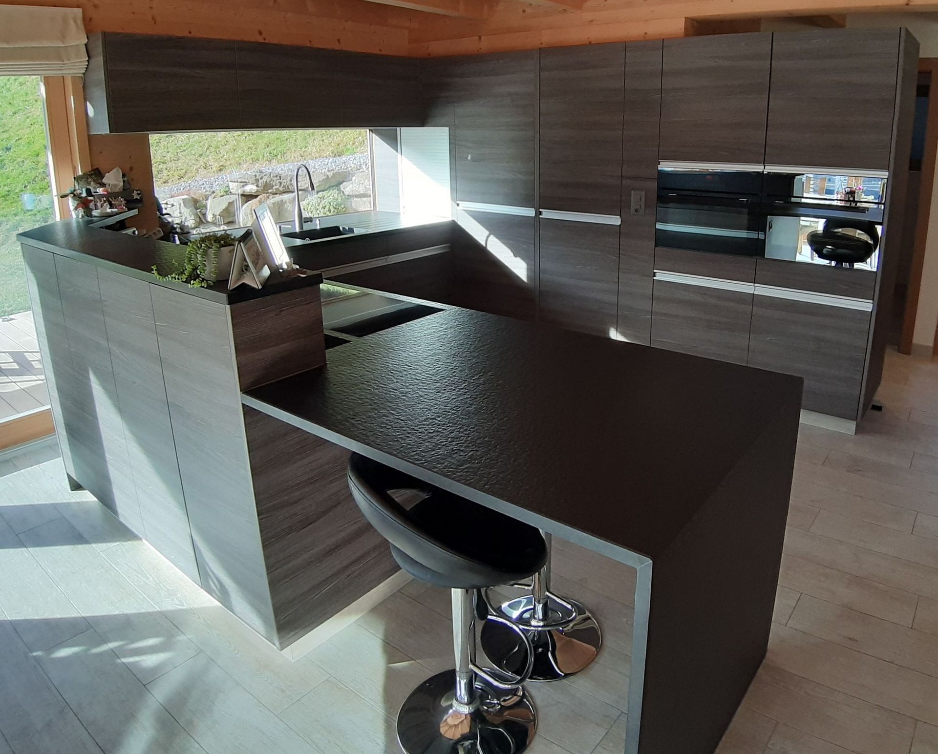 Cuisine en stratifiée gris imitation bois, plan de travail en granit noir absolu, poignées profilées en alu, hotte de ventilation intégrée dans le plan de travail, store déroulant et porte secrète qui donne sur le cellier!