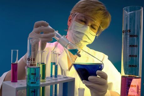 Científico con bata de laboratorio y mascarilla prueba un líquido azul con material de vidrio entre coloridos vasos de precipitados y probetas graduadas.