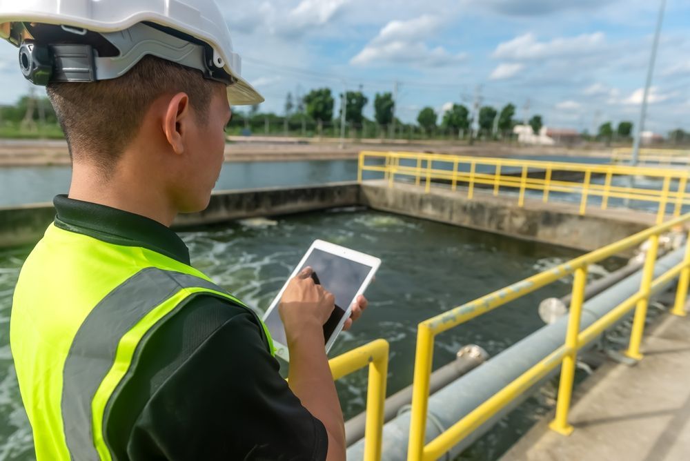 Un trabajador utiliza una tableta junto a un tanque de tratamiento de aguas residuales con barandillas de seguridad amarillas.