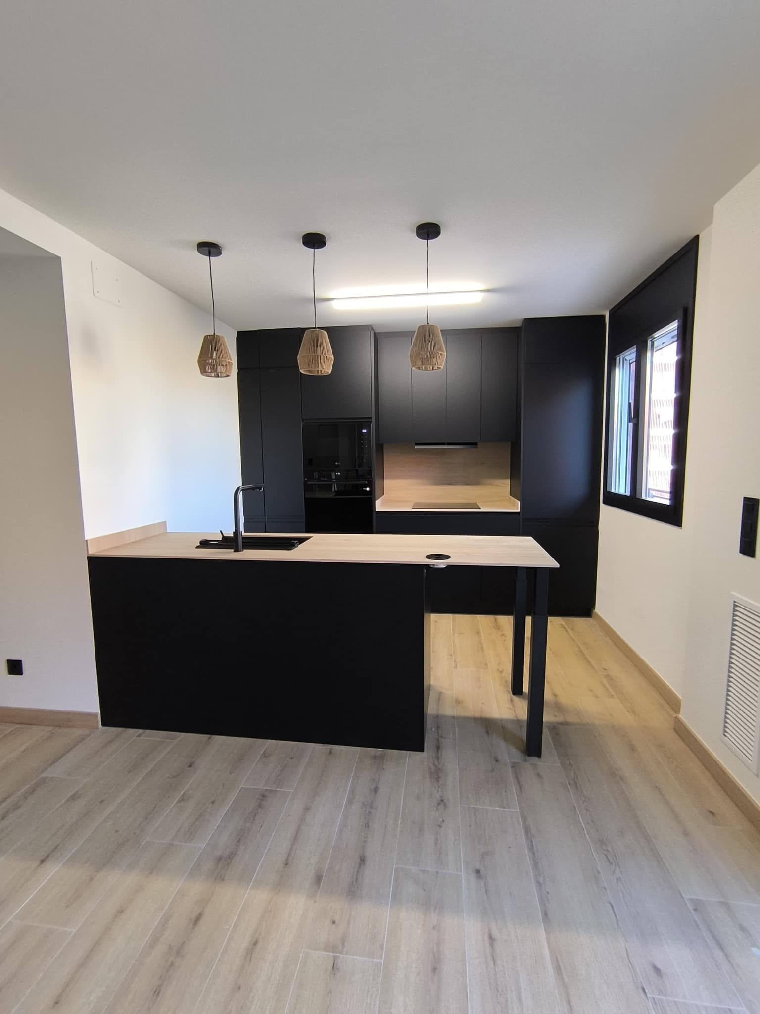 Cocina moderna negra con detalles de madera; una isla de cocina y luces colgantes.