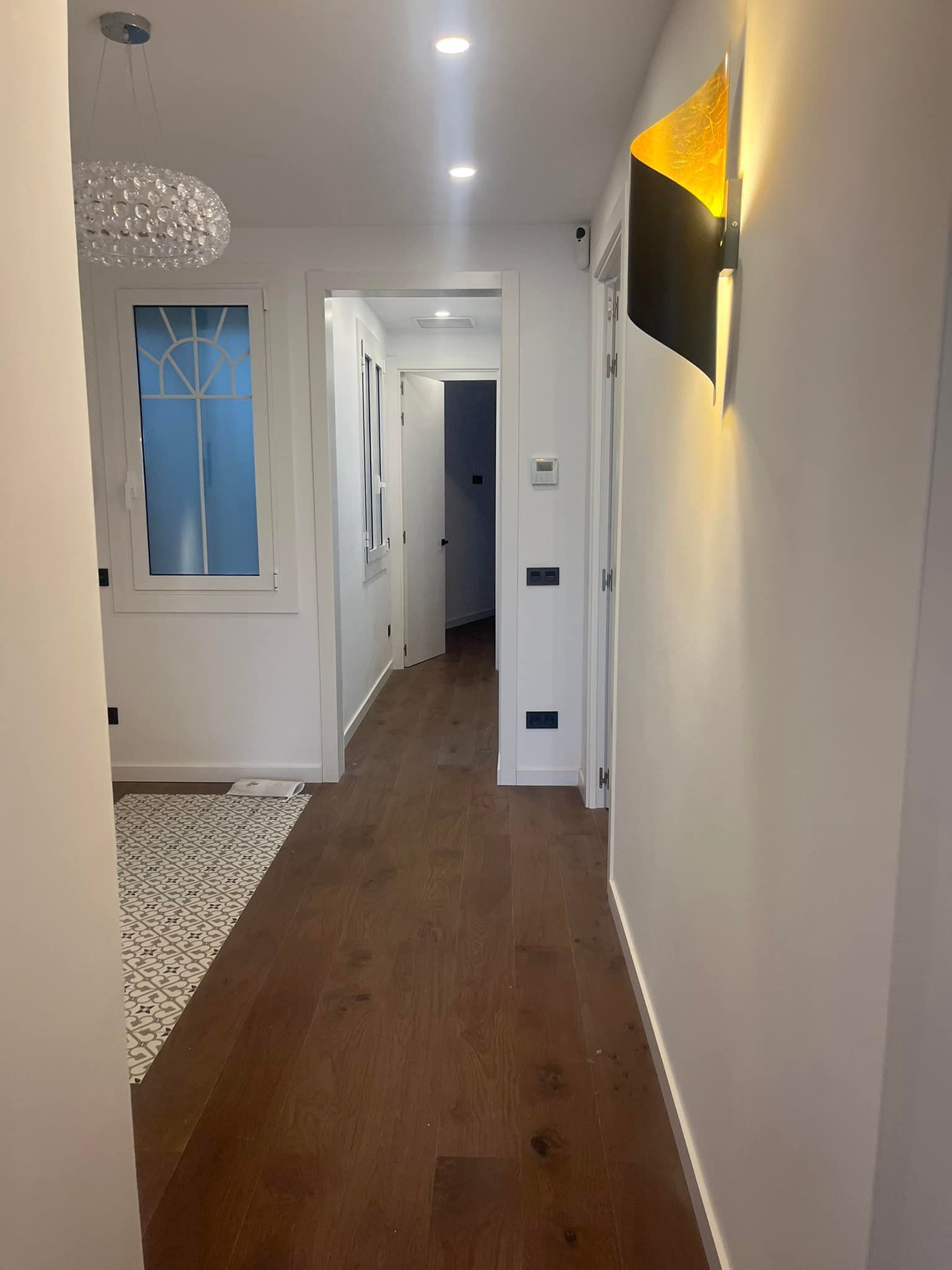 Pasillo estrecho con piso de madera, paredes blancas, aplique de pared dorado moderno y puertas abiertas.