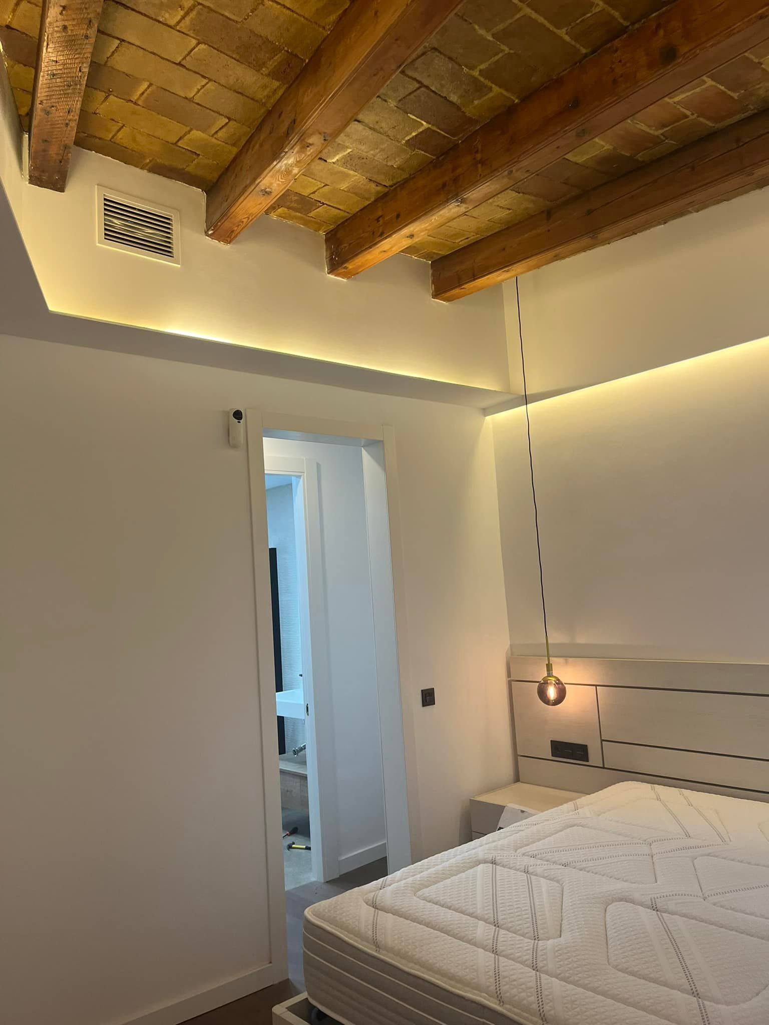 Dormitorio con vigas de madera en el techo, iluminación empotrada y cama con cabecero blanco.