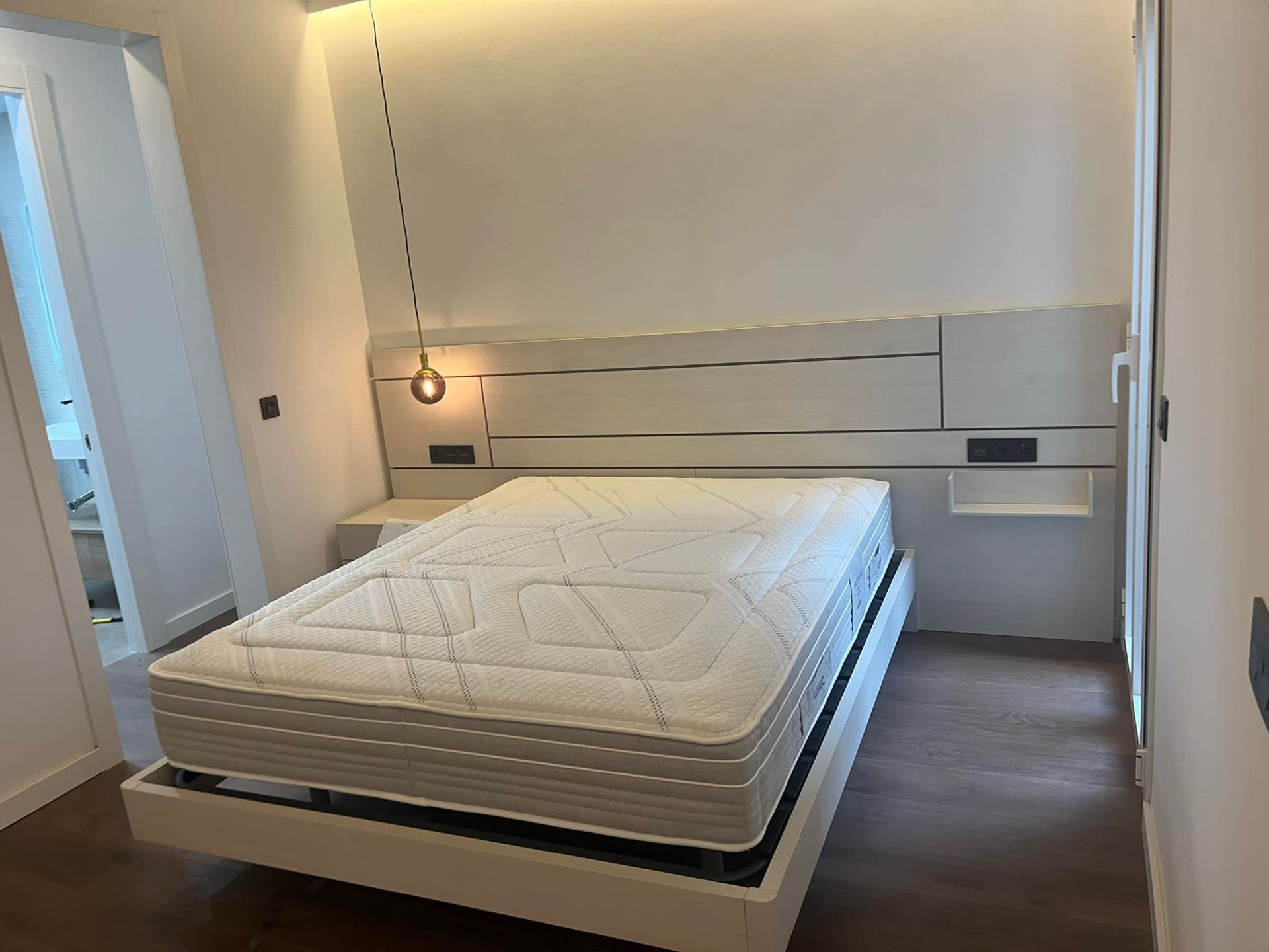 Dormitorio con cabecero blanco, colchón y mesita de noche flotante.