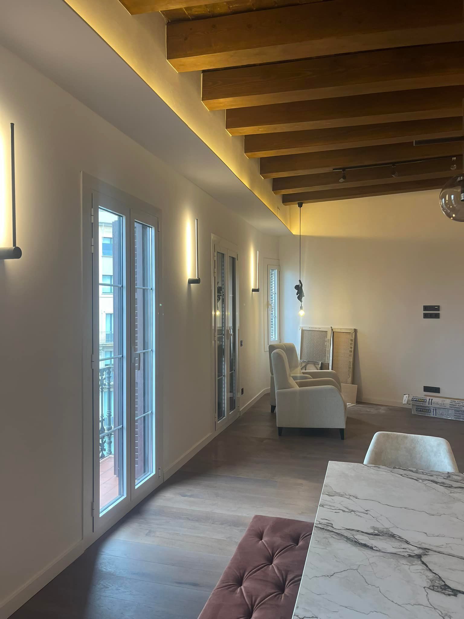 Una habitación larga y luminosa con una mesa de mármol, paredes blancas, vigas de madera en el techo y ventanas altas.