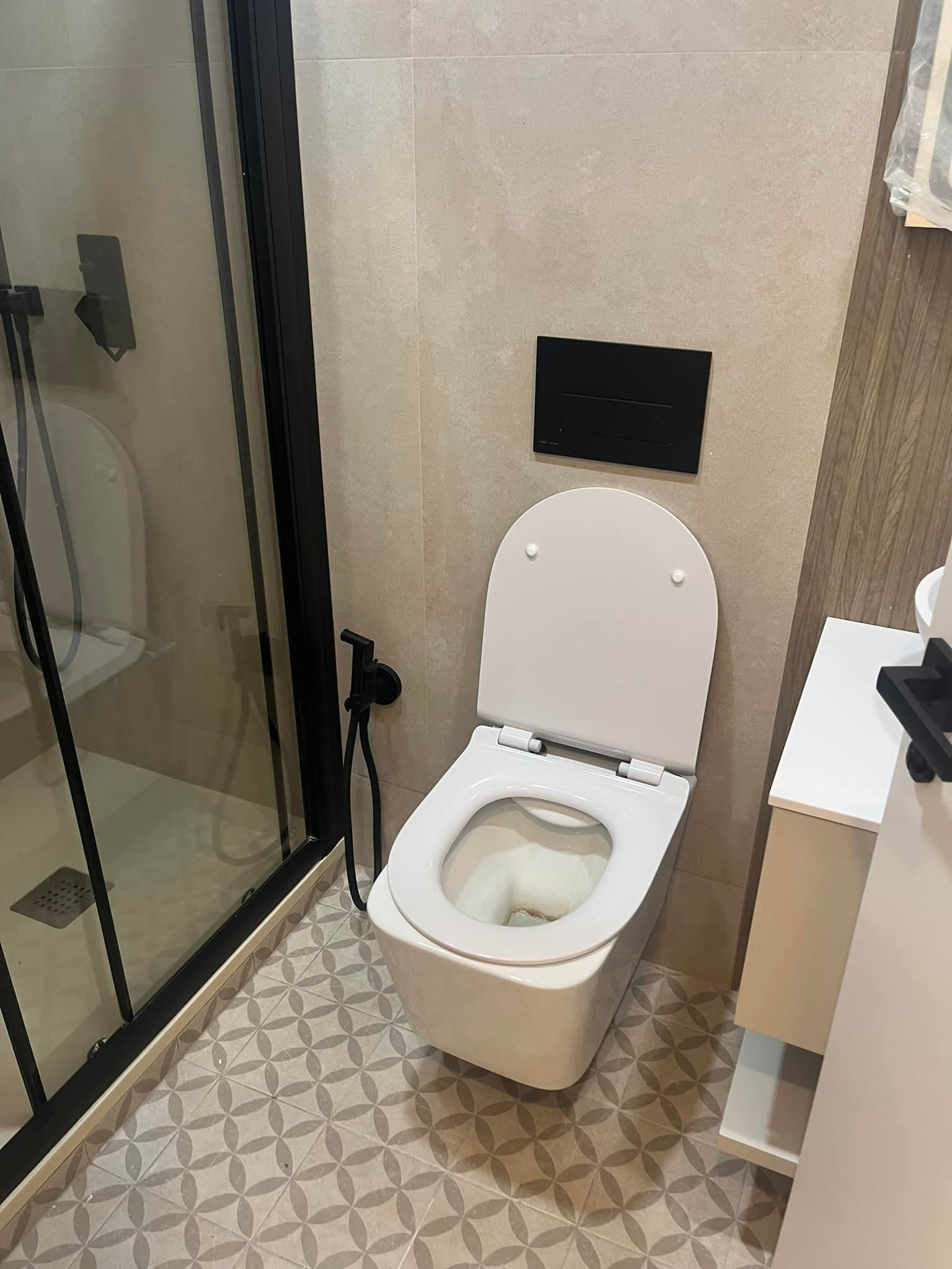 Inodoro con tapa abierta en baño moderno con marco de ducha negro, paredes beige y piso de baldosas estampadas.