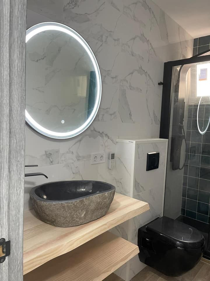 Baño moderno con inodoro negro, lavabo de piedra, estante de madera y espejo iluminado.