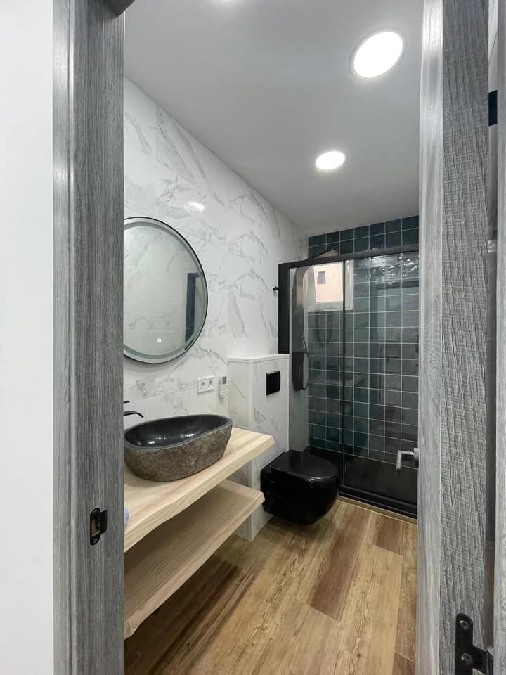 Baño con lavabo de piedra, espejo redondo, pared de mármol e inodoro y ducha de color negro.