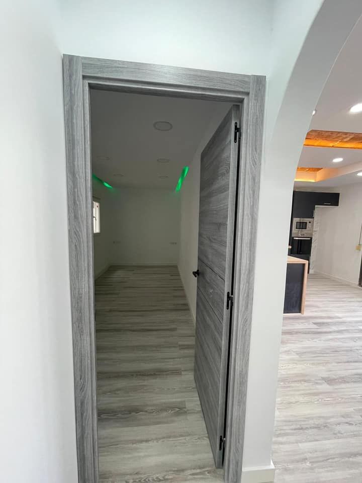 La puerta y el marco grises se abren a una habitación con piso a juego y paredes blancas; la puerta conduce a la cocina.