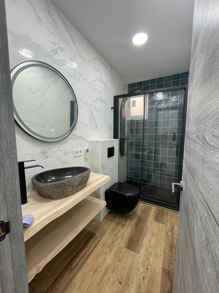 Baño moderno con lavabo de piedra, inodoro negro, ducha, piso de madera y paredes revestidas de mármol y azulejos verde azulado.