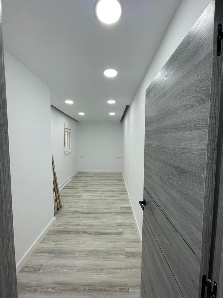 Pasillo estrecho y vacío con suelo de imitación madera clara y paredes blancas, iluminado por luces empotradas en el techo. Una puerta gris está abierta.